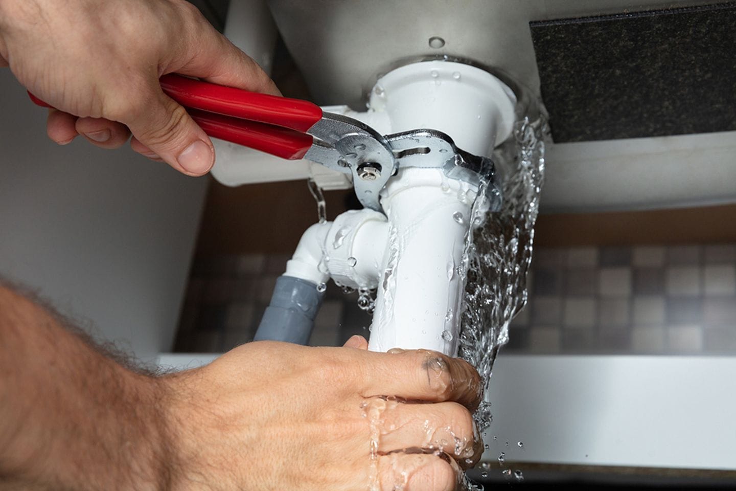 Person using pliers to fix a leaking pipe under a sink, with water spilling from the joint onto hands.