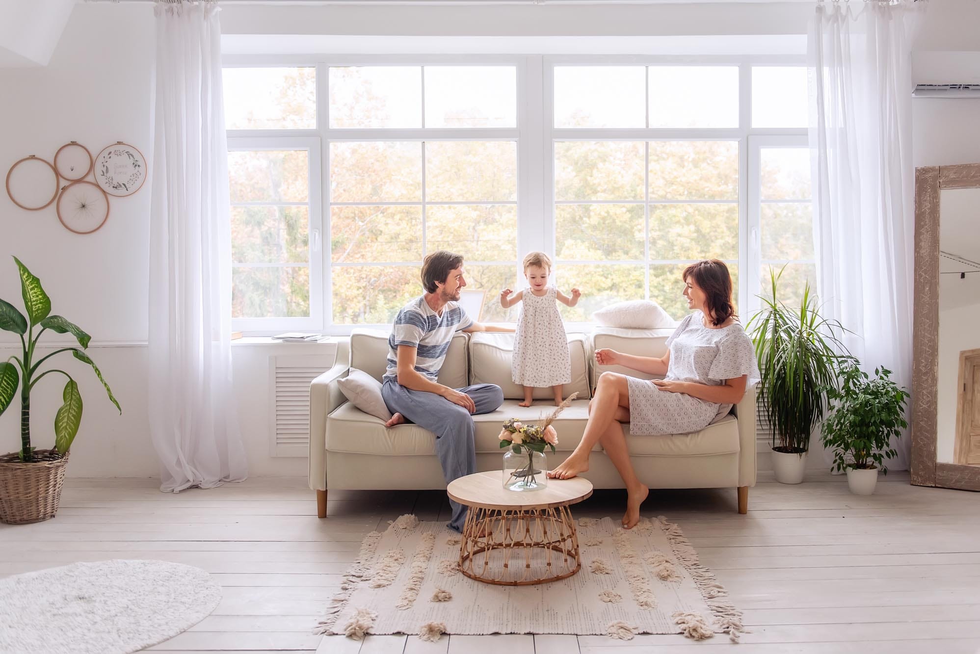 Family enjoying time together in a bright living room, with a child standing on a sofa, parents engaged in conversation, and a coffee table with flowers.