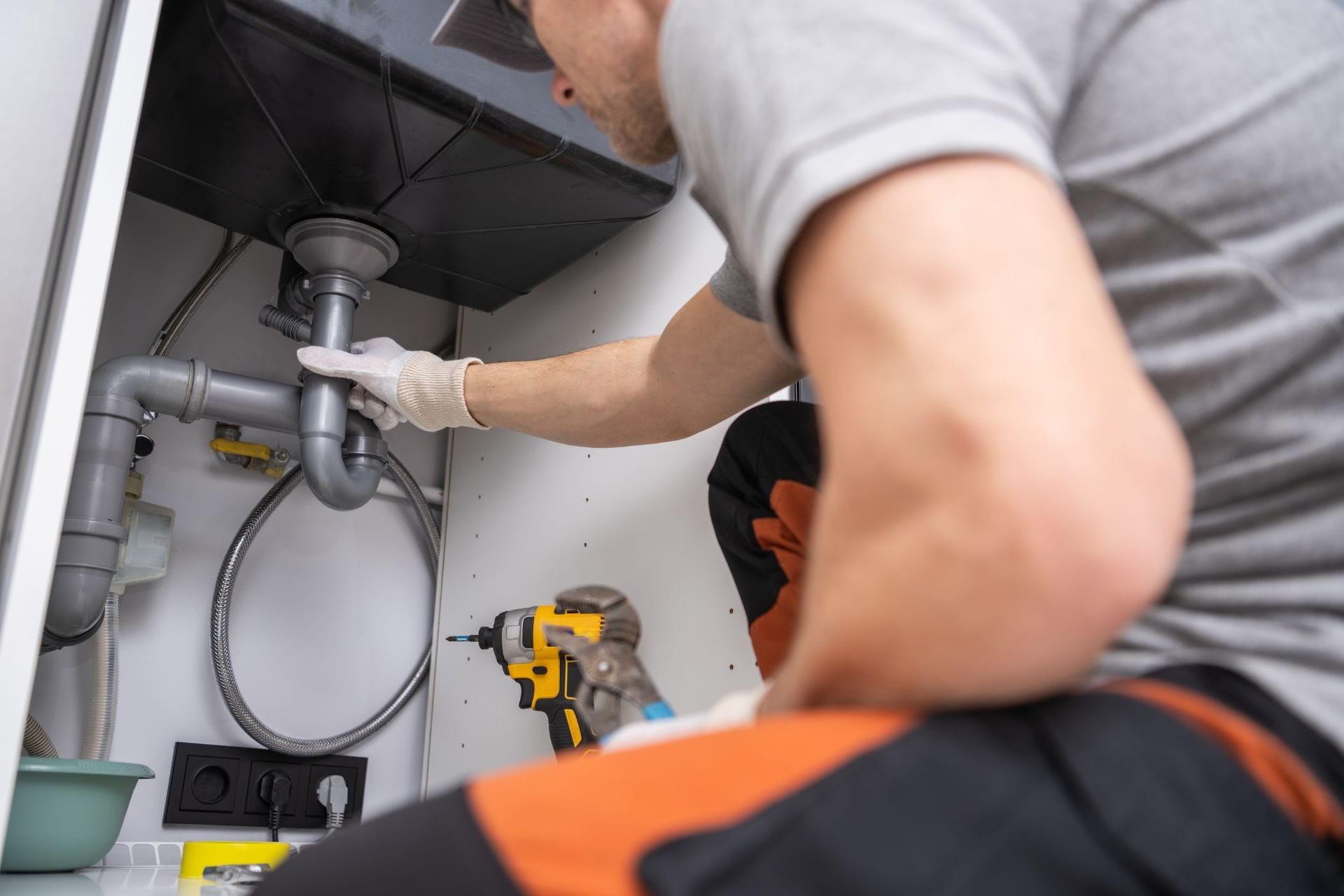 A plumber repairs pipes under a kitchen sink, working with tools like a drill and wrench.
