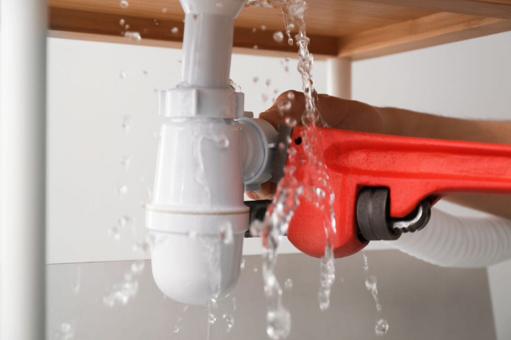Water leaking from a white plumbing pipe under a sink, with a person using a red wrench for repairs, illustrating urgent plumbing issues related to leak detection and water line repair services in Atlanta, GA.
