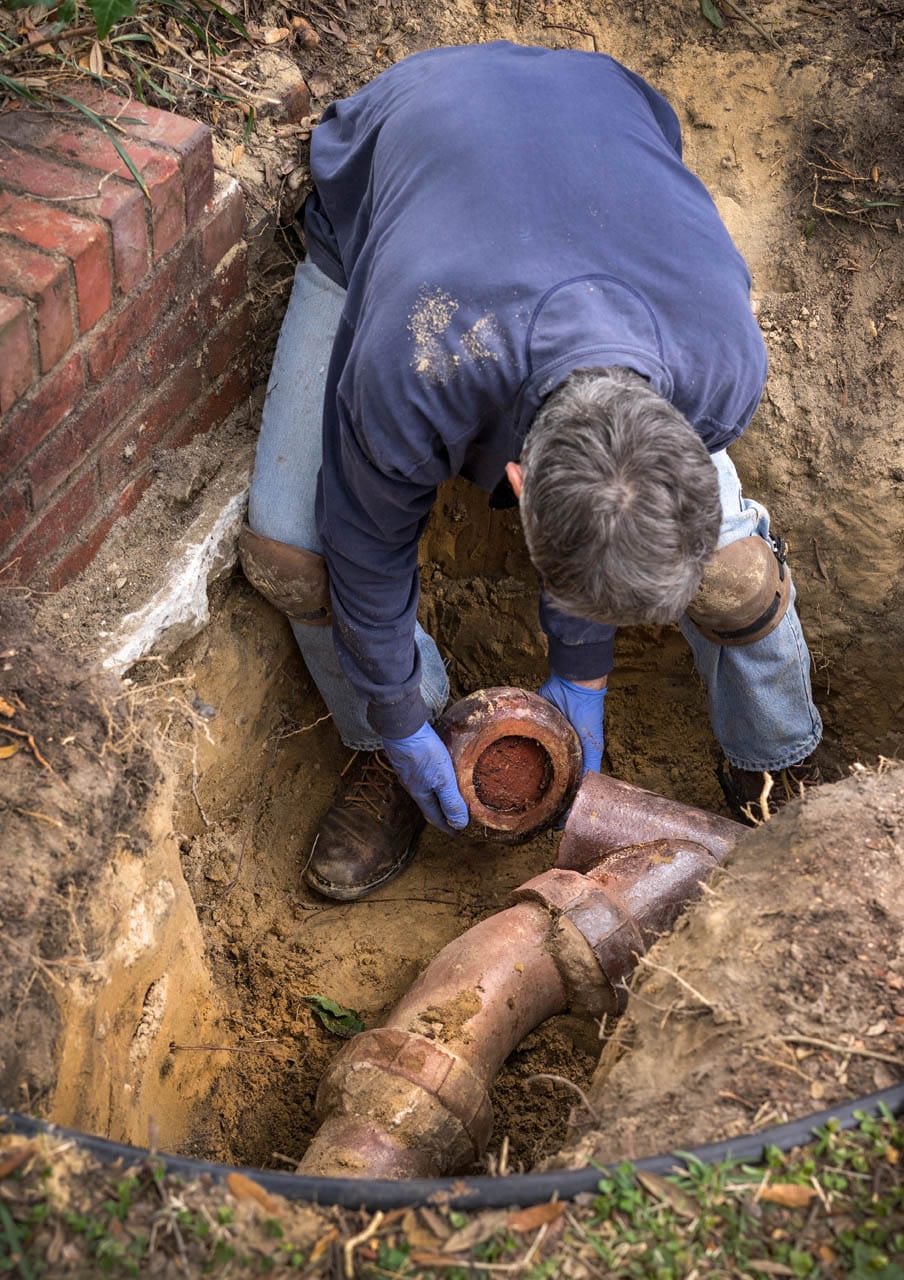 Plumber repairing sewer line, handling pipe in excavation, surrounded by dirt and grass, illustrating professional sewer line repair services in Northwest Georgia.