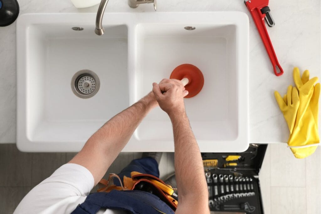 Person using a plunger in a double sink to address potential drain clogs, with yellow rubber gloves and plumbing tools visible, emphasizing practical drain maintenance.
