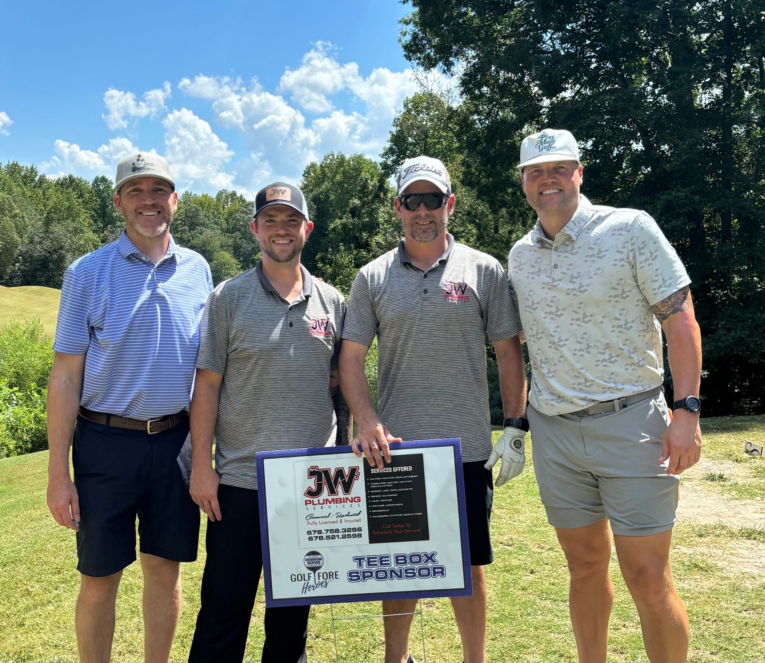 Four men standing on a golf course, wearing JW Plumbing Pros branded shirts and hats, holding a sign that promotes their plumbing services and indicates they are tee box sponsors for a charity event.