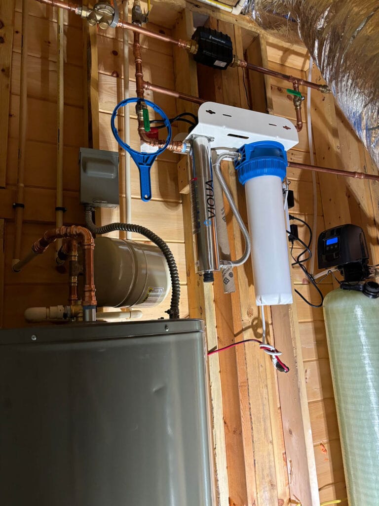 Water filtration system with pipes, a pressure tank, and a filter in a wooden enclosure. Various connectors and valves are attached to the plumbing arrangement.