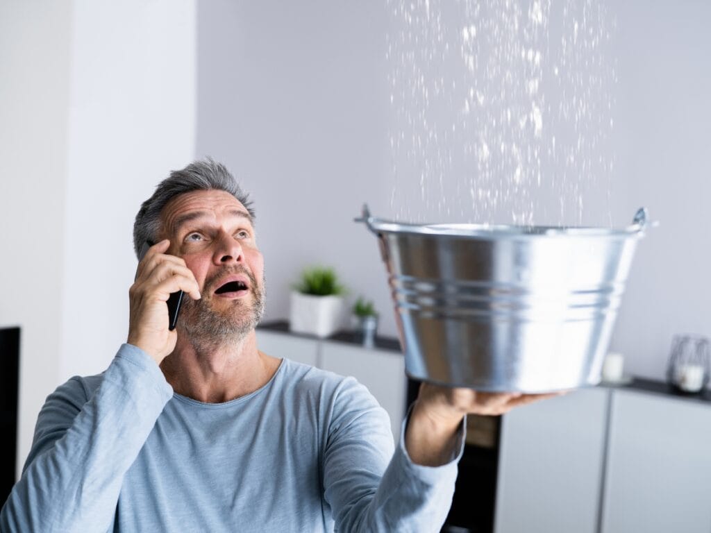Man holding a metal bucket to catch water leaking from the ceiling while talking on a phone, illustrating the urgency of addressing hidden water leaks in homes.