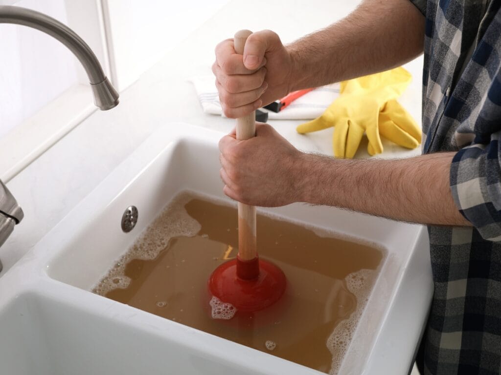 Person using a red plunger to clear a clogged sink filled with murky water, emphasizing DIY drain cleaning solutions for recurring clogs.