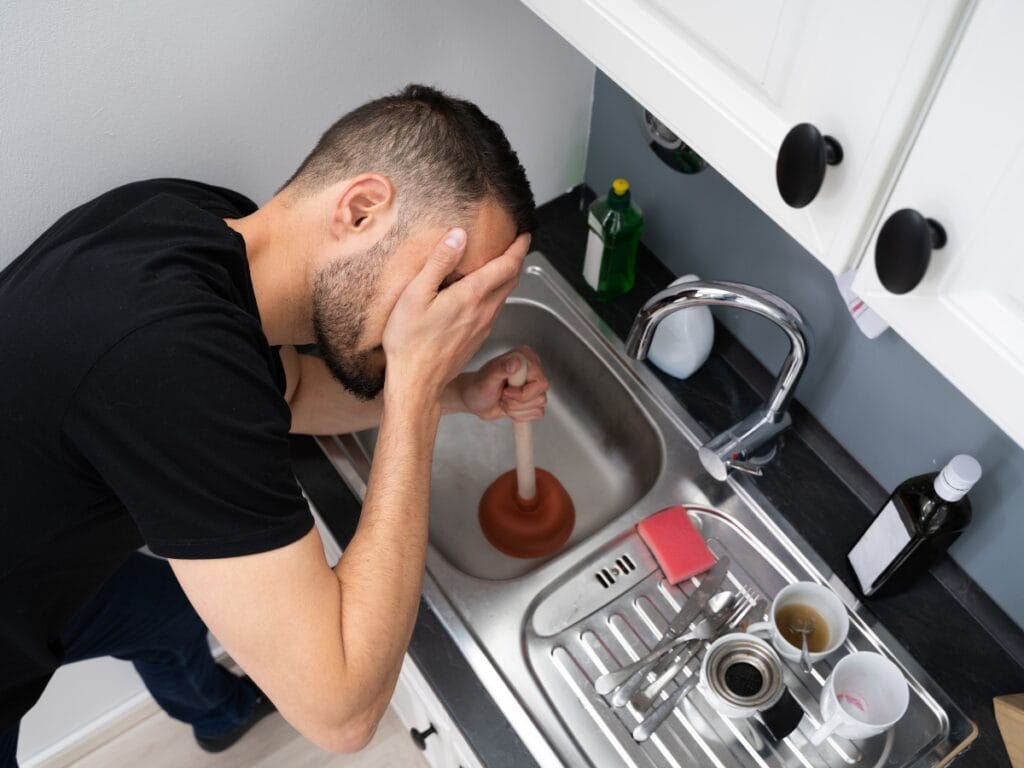 Man using a plunger in a kitchen sink, showing frustration with a recurring drain clog, amidst dirty dishes and cleaning supplies, illustrating common plumbing problems faced by homeowners.