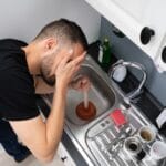 Man using a plunger in a kitchen sink, expressing frustration over recurring drain clogs, related to plumbing issues discussed by JW Plumbing Pros.