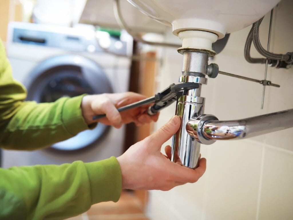 Person using adjustable wrench to repair plumbing under a sink, focusing on plumbing issues common in older homes, such as corroded pipes and fittings.