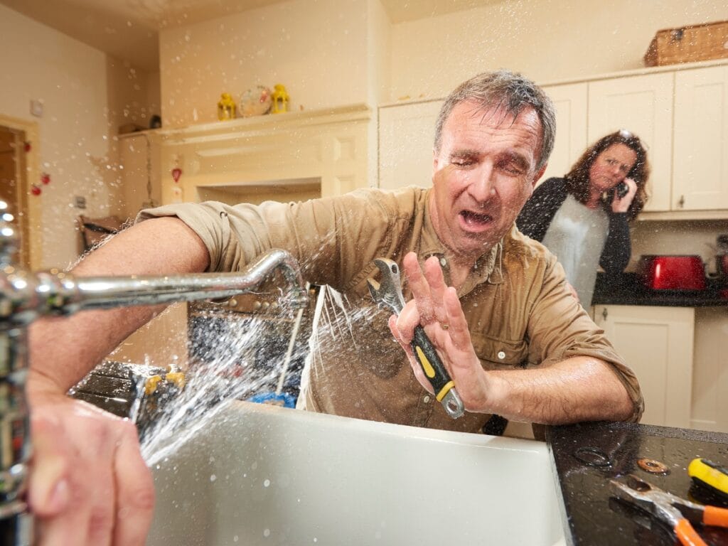 Man struggling with a leaky kitchen sink, water spraying, while a woman on the phone looks concerned, illustrating common plumbing problems in old houses.