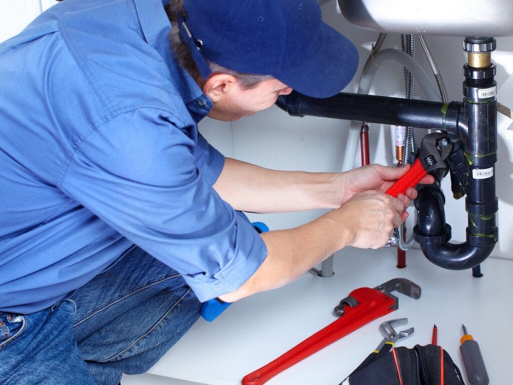 Plumber using a red wrench to repair pipes under a sink, surrounded by plumbing tools, illustrating maintenance for common plumbing issues in older homes.