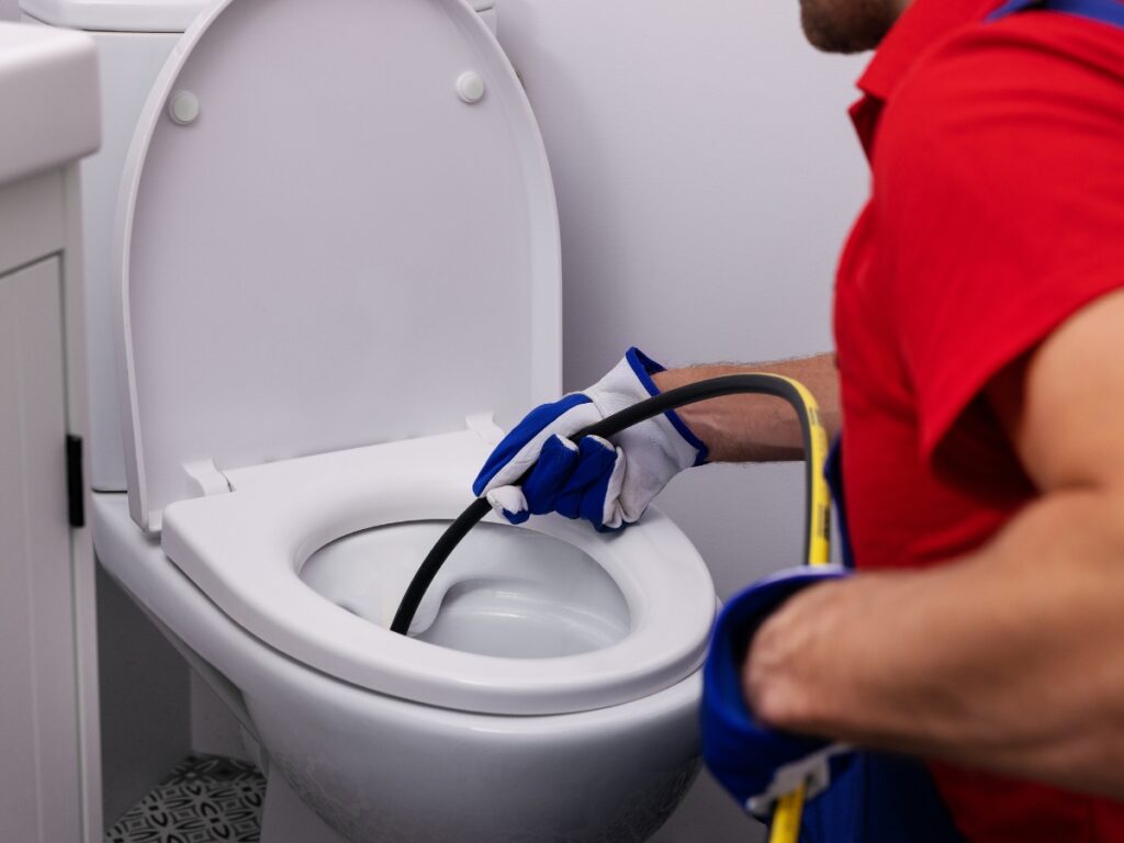 A person wearing gloves uses a plumbing snake to clear a clogged toilet in a bathroom.