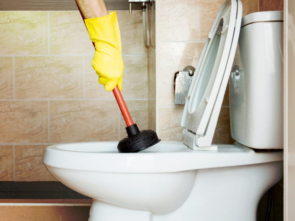 A person wearing a yellow rubber glove uses a plunger to fix a clogged toilet in a bathroom with beige tiled walls.