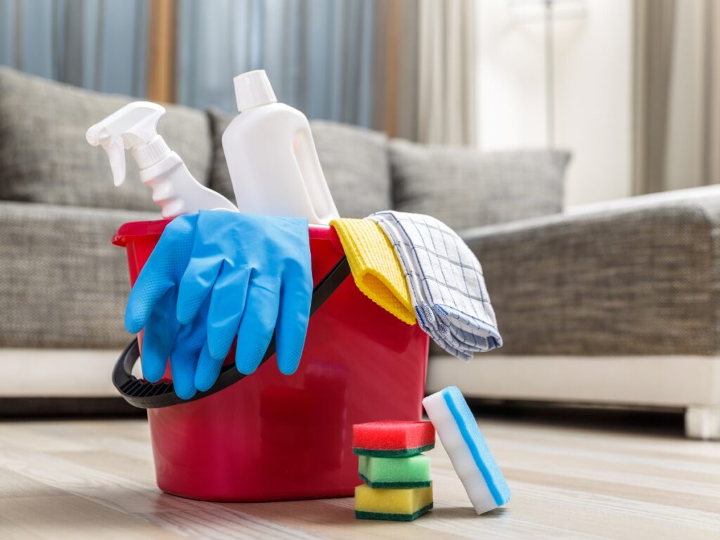 A red bucket filled with cleaning supplies, perfect for tackling any mess—even a clogged toilet—sits on the floor with sponges nearby and a sofa in the background.