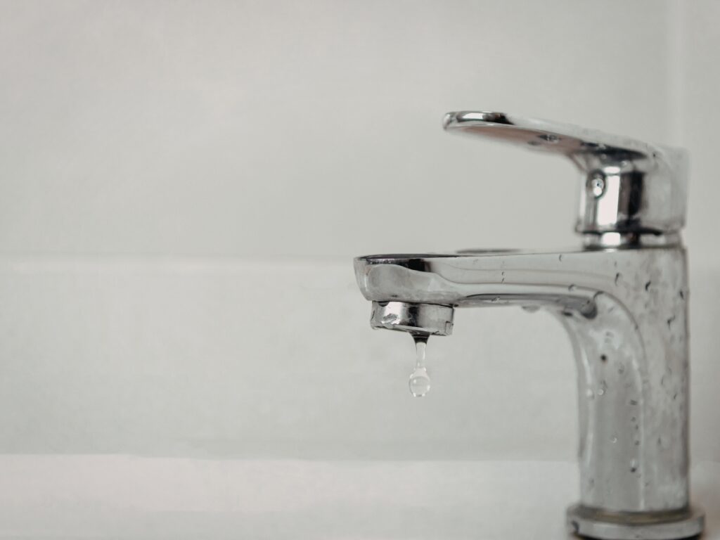 Close-up of a chrome faucet with a single water droplet falling from the spout, capturing the moment of a dripping faucet against a plain, light-colored background.