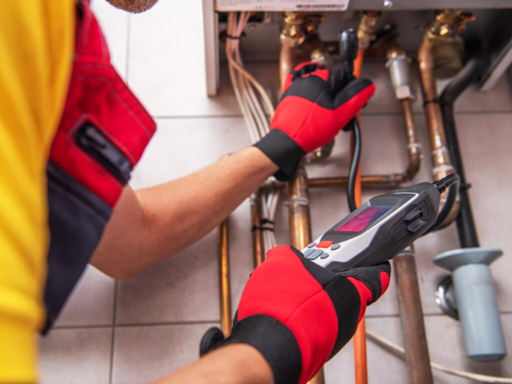 A worker wearing red gloves uses a diagnostic tool to inspect pipes and connections beneath a boiler, looking for signs of issues such as a dripping faucet.