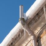 Close-up of a building&rsquo;s roof edge with snow and icicles, showing a drainpipe system designed to minimize damage from frozen burst pipes against a clear blue sky.