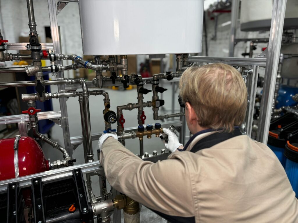Wearing work gloves, a person adjusts valves on a complex system of pipes and gauges in an industrial setting to minimize damage from frozen burst pipes.
