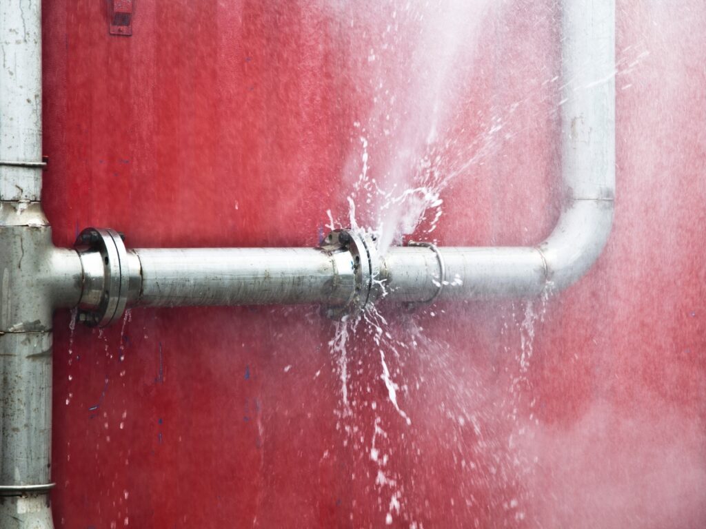 Water is spraying from a frozen burst pipe in a metal line mounted on a red wall. Take action quickly to minimize damage.