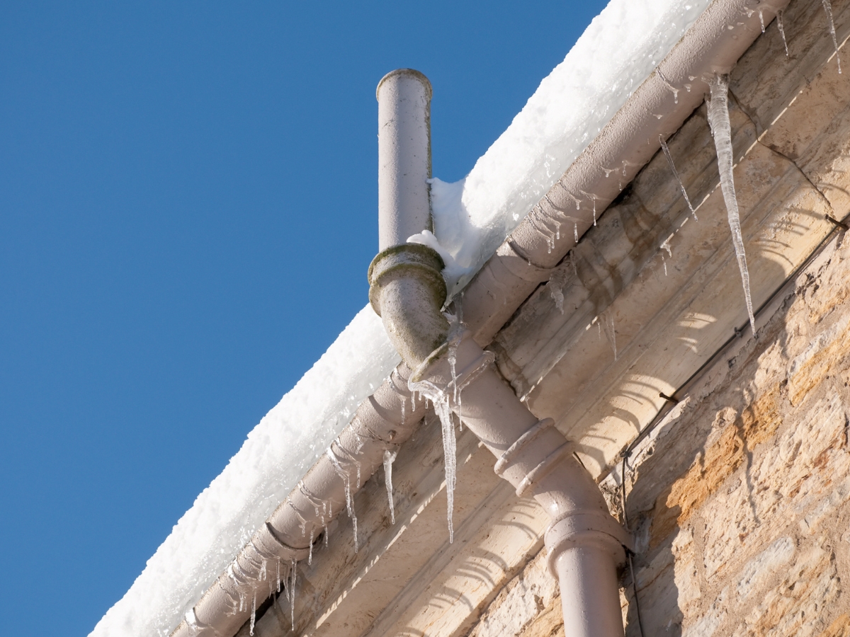 Close-up of a building&rsquo;s roof edge with snow and icicles, showing a drainpipe system designed to minimize damage from frozen burst pipes against a clear blue sky.