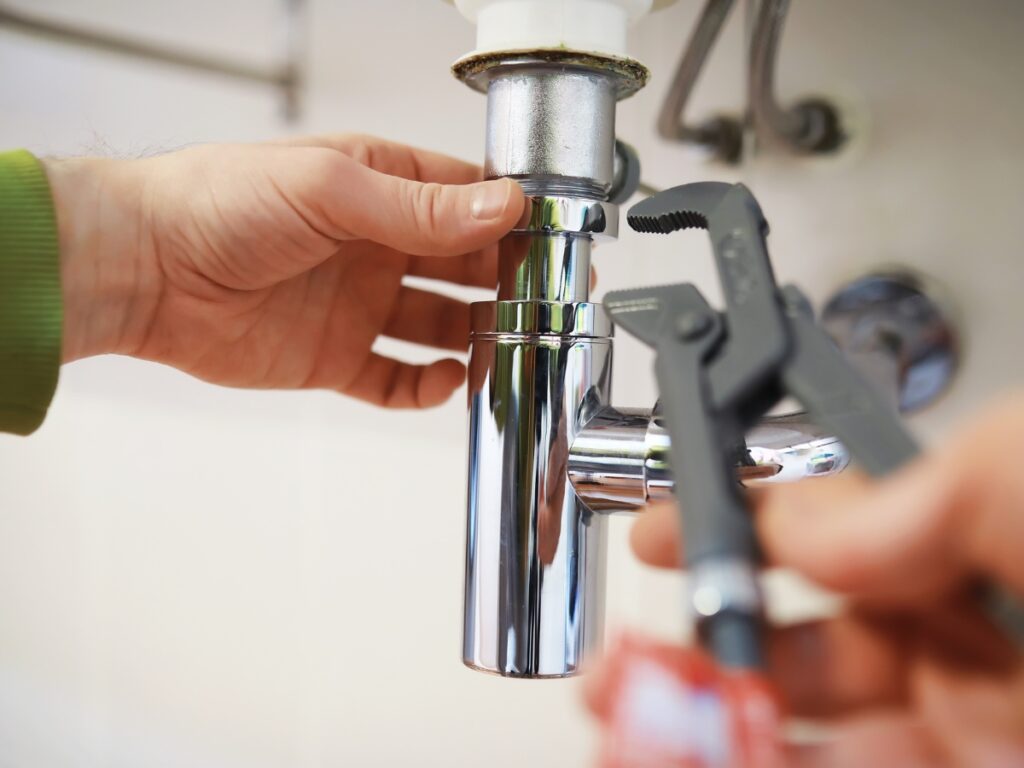A person uses a wrench to tighten a chrome pipe under a sink, performing plumbing maintenance to prevent costly repairs.