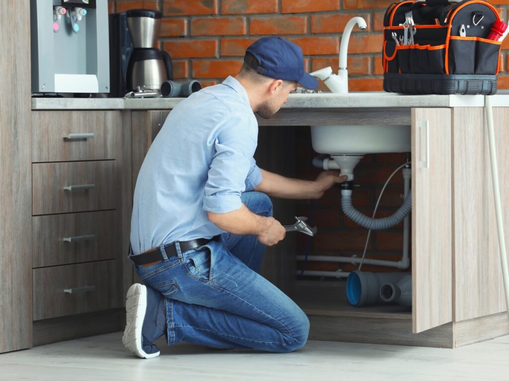 Plumber inspecting and repairing kitchen sink plumbing under cabinet, showcasing maintenance and DIY solutions for homeowners.