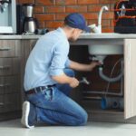 A man kneels under a kitchen sink, holding a wrench and working on plumbing maintenance. A tool bag sits on the countertop above him, highlighting the importance of regular upkeep to prevent costly repairs.
