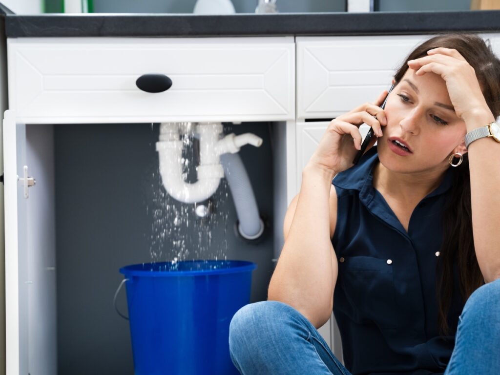 A woman sits on the floor, talking on the phone and looking stressed as water leaks from a pipe under the sink into a blue bucket&mdash;reminding us how plumbing maintenance can help prevent costly repairs.