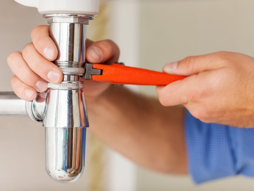 A person uses a wrench to tighten or loosen a metal pipe under a sink, performing plumbing maintenance to prevent costly repairs.