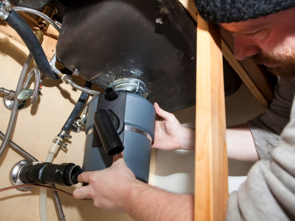 Person installing a garbage disposal under a kitchen sink, showcasing plumbing components and maintenance practices related to disposal repair and replacement.
