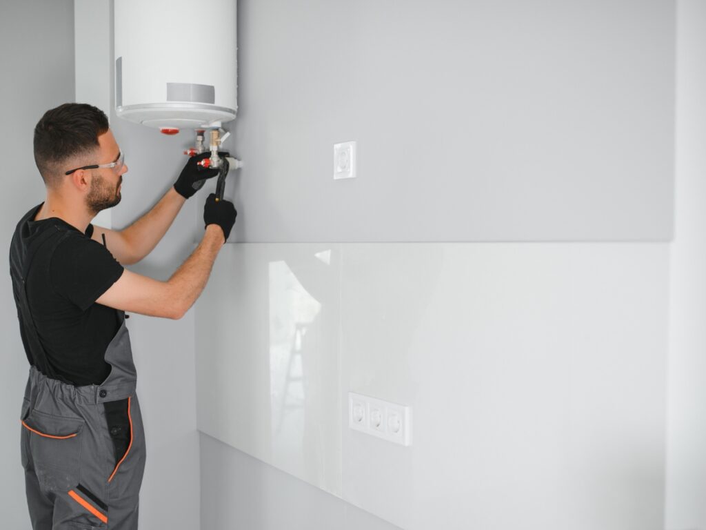 A person wearing work overalls and gloves uses a tool to adjust or install a wall-mounted water heater in a modern, gray room, possibly addressing issues with water heaters not heating.