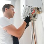 A person wearing safety glasses and gloves is repairing or inspecting a wall-mounted gas boiler with exposed pipes and valves, possibly diagnosing issues such as water heaters not heating.