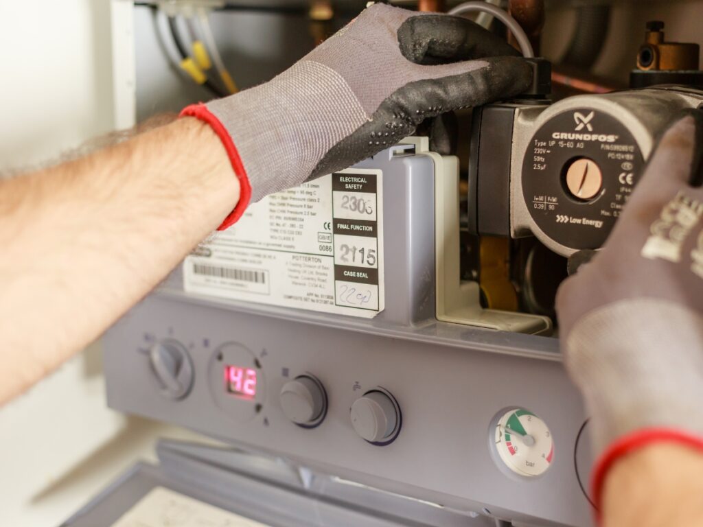 A person wearing gloves adjusts a component inside a gas boiler, with a digital display showing 42°C—possibly troubleshooting why water heaters are not heating, as various gauges and labels are visible.