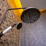 Close-up of a modern showerhead releasing gentle streams of water, ideal for low pressure systems, with a smaller handheld showerhead beside it and a tiled wall in the background.