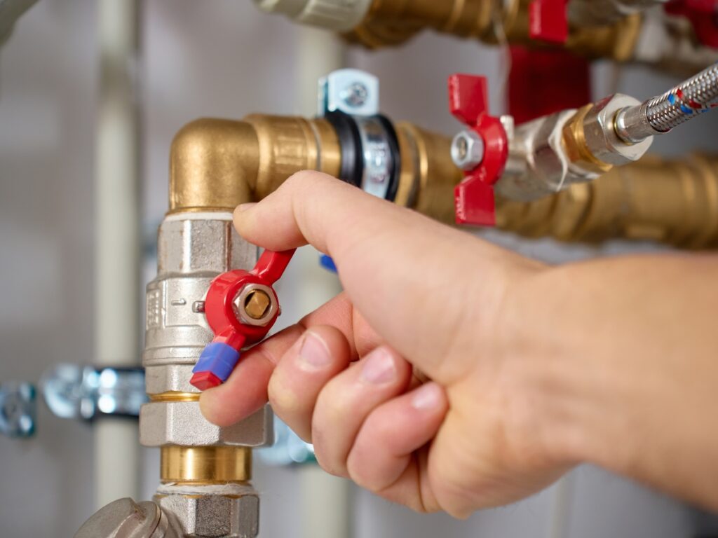 A person’s hand turns a red valve handle on a metal plumbing pipe connected to other pipes and fittings, adjusting for low pressure.