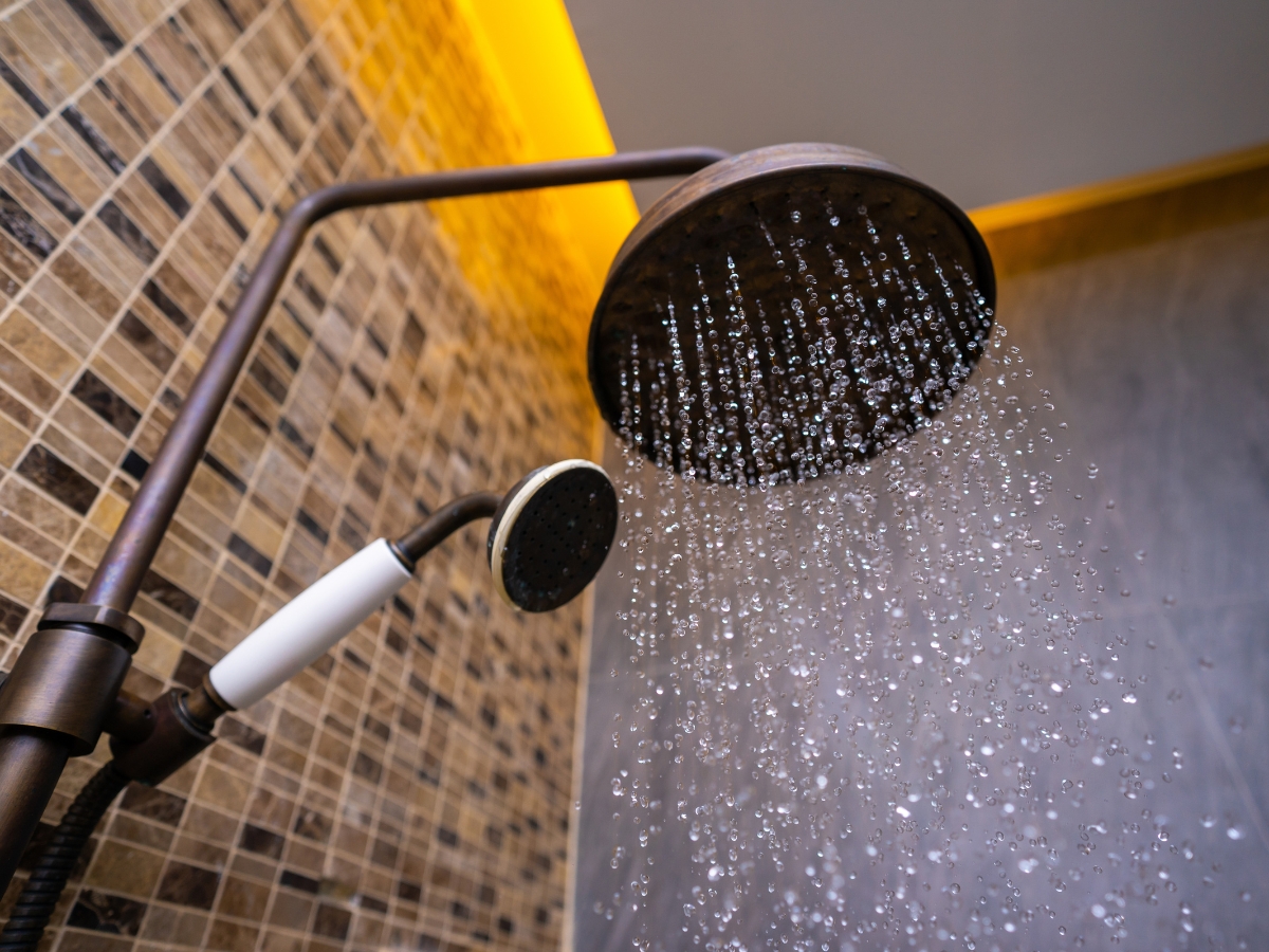 Close-up of a modern showerhead releasing gentle streams of water, ideal for low pressure systems, with a smaller handheld showerhead beside it and a tiled wall in the background.