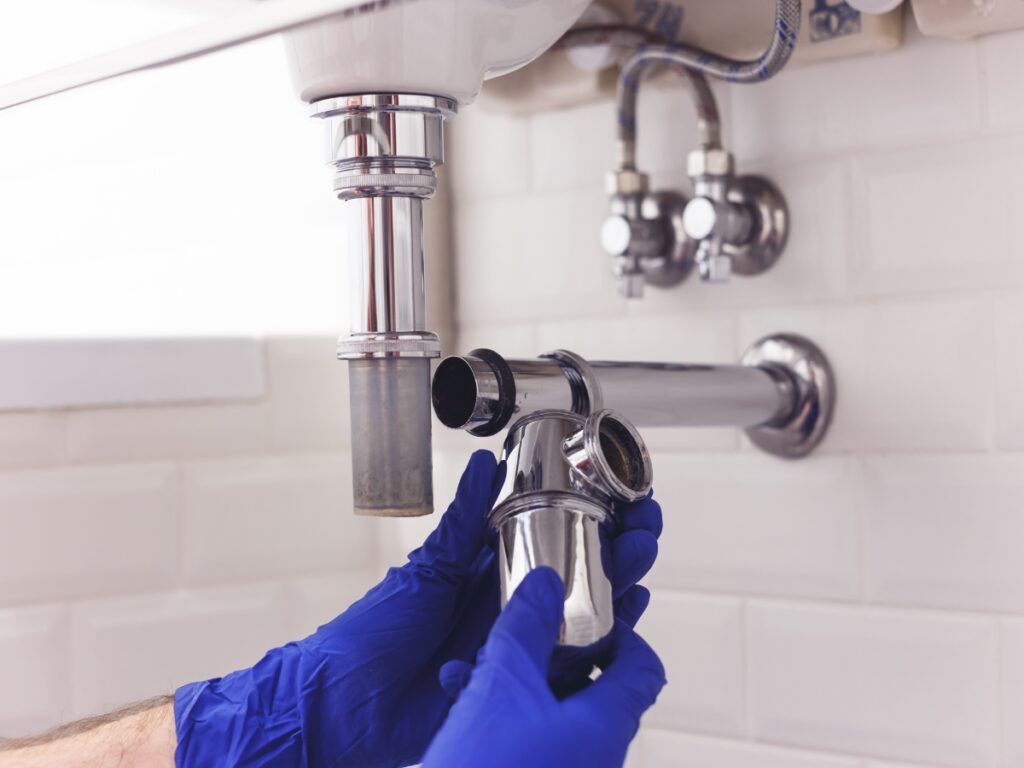Person wearing blue gloves repairing or inspecting a metal sink drain trap under a bathroom sink with a white tiled wall in the background, following tips from a slab leak guide.
