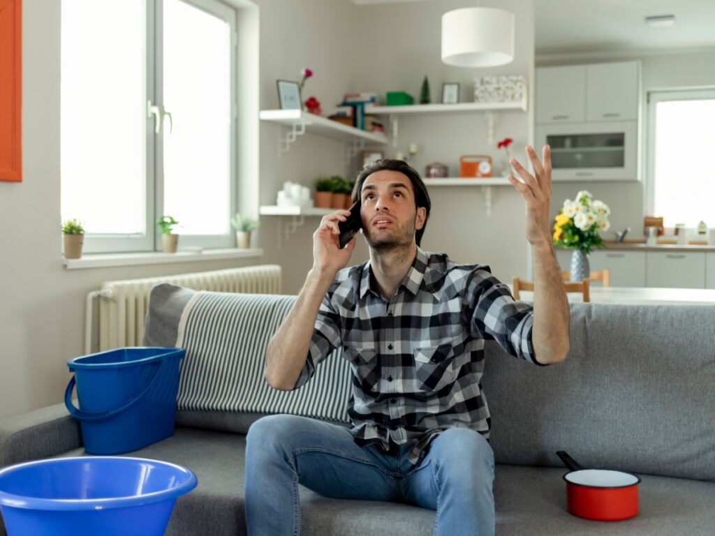 A man sits on a couch talking on the phone, looking up with concern. Buckets and a pot on the floor suggest he is dealing with a water leak in his home, possibly searching for a slab leak guide to fix the issue.