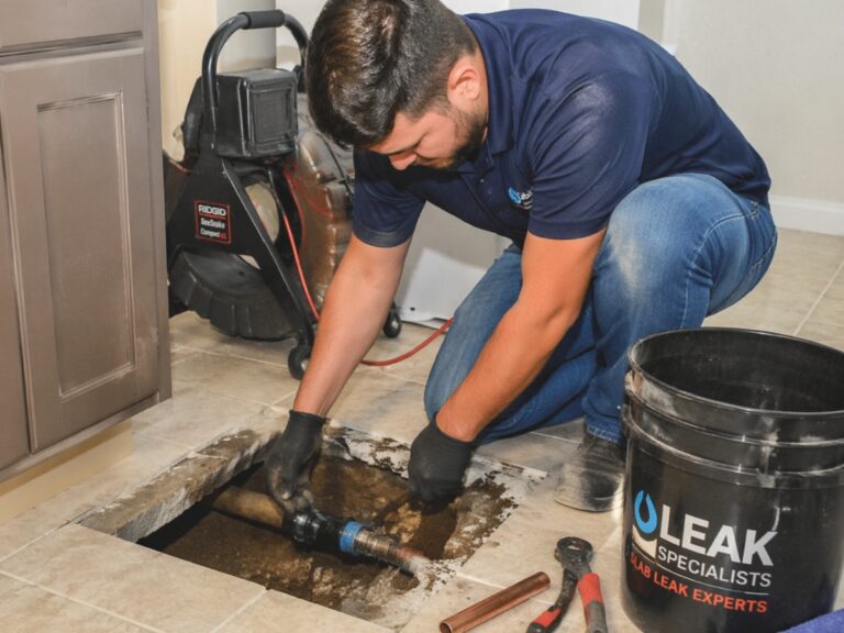 A plumber repairs a pipe under a kitchen floor, surrounded by tools and a bucket labeled “Leak Specialists,” following tips from a slab leak guide.