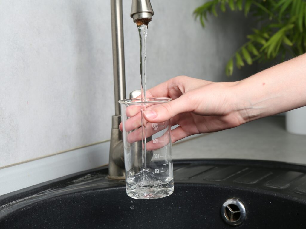 A person holds a glass beaker under a running faucet, filling it with water at a sink—an ideal setting to demonstrate water filtration. A green plant is visible in the background.