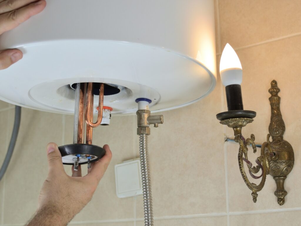 A person is repairing or inspecting the heating element at the bottom of a wall-mounted water heater with integrated water filtration, near a lit wall sconce.