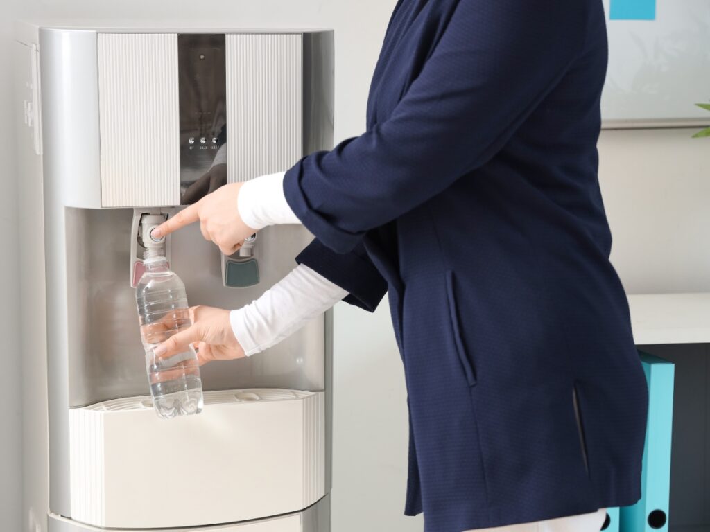 A person in a dark blazer fills a plastic water bottle at a water dispenser equipped with advanced water filtration.