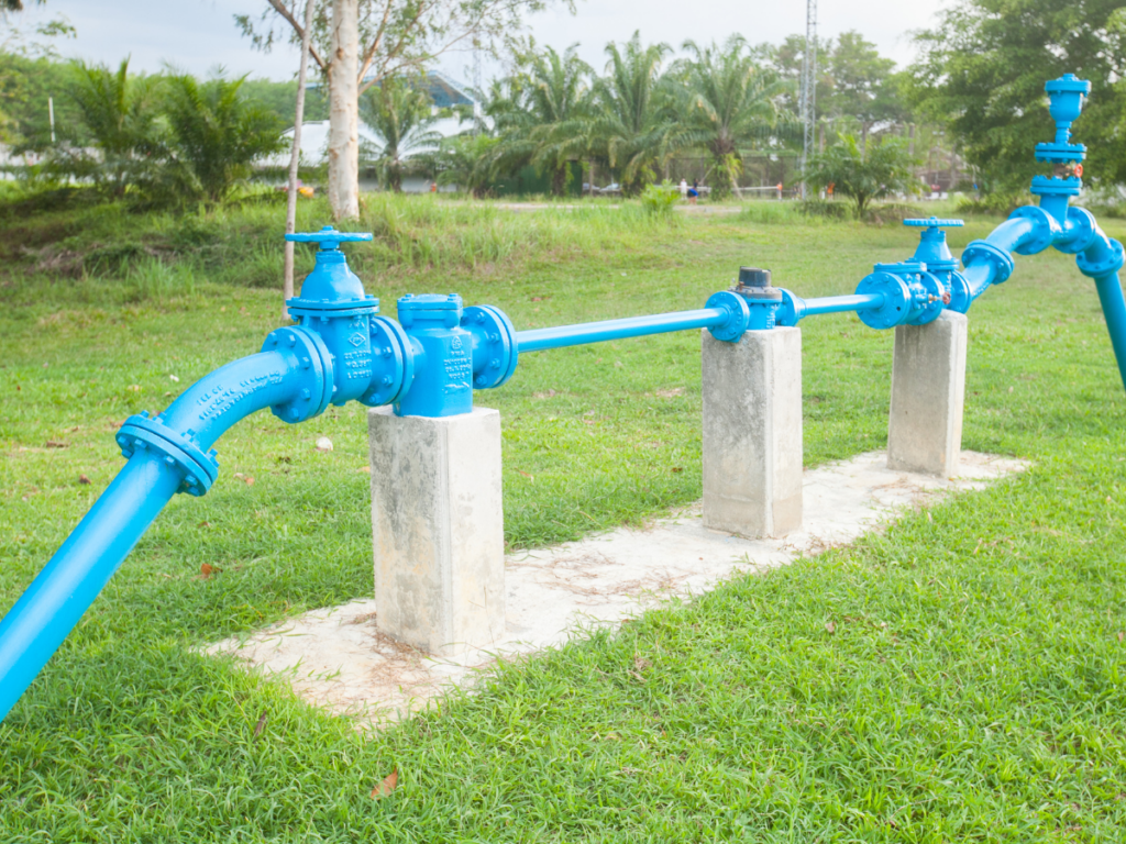 A blue metal pipeline system with valves and a backflow prevention device, supported by three concrete posts, is installed on a grassy area outdoors.