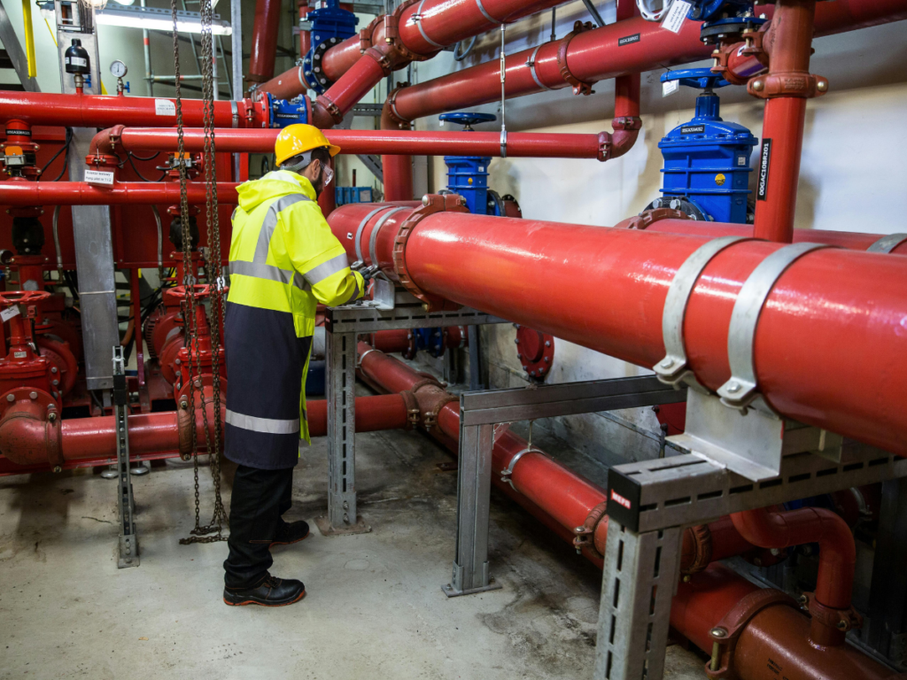 A worker in safety gear inspects large red industrial pipes and valves, ensuring proper backflow prevention inside a mechanical room.
