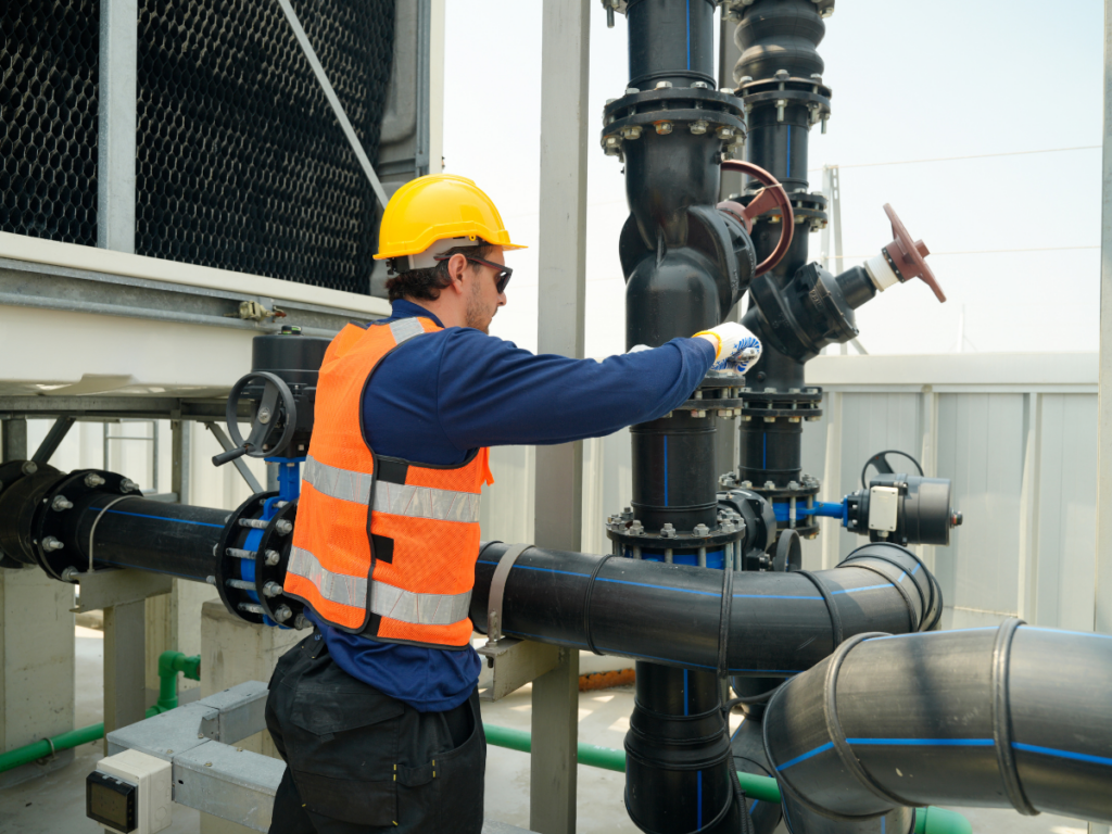 Worker wearing a yellow helmet and orange safety vest adjusts a large industrial valve with backflow prevention on black piping in an outdoor facility.