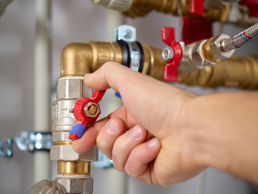 A person’s hand turning a red shut-off valve on a metal water pipe with backflow prevention, connected to a plumbing system.