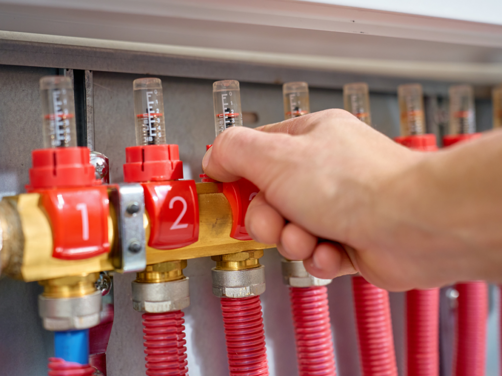 A hand adjusts a red valve on a manifold with numbered dials and red flexible pipes in a plumbing or heating system, ensuring proper backflow prevention.