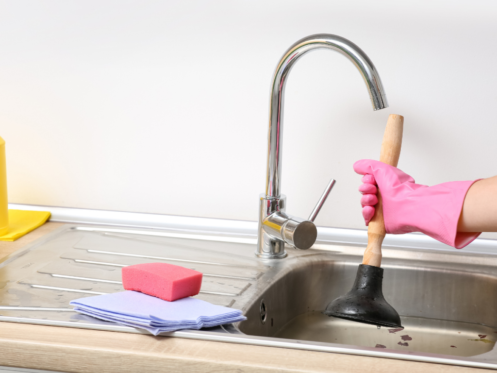 A person wearing a pink glove uses a plunger in the kitchen sink, demonstrating effective methods for preventing clogs; a pink sponge and blue cloths are on the countertop nearby.