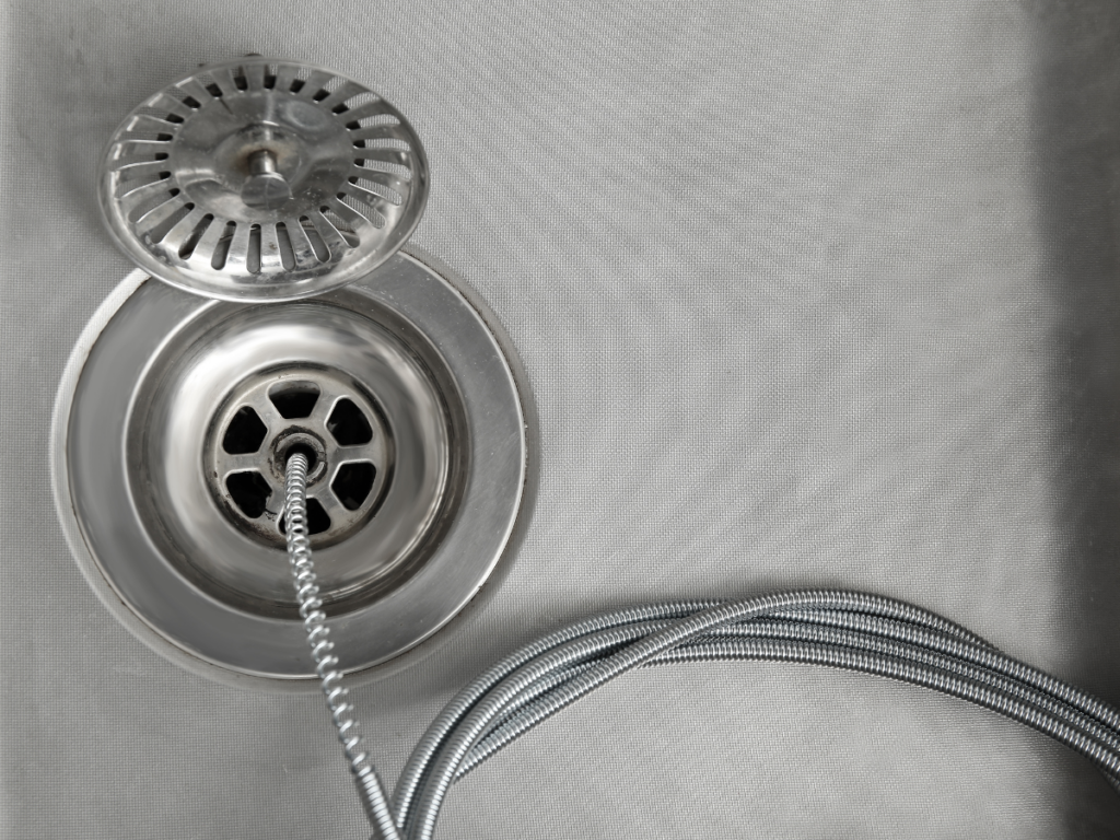 Close-up of a metal sink drain with a flexible hose inserted, and the drain cover resting on the edge—an effective method for preventing clogs during cleaning.