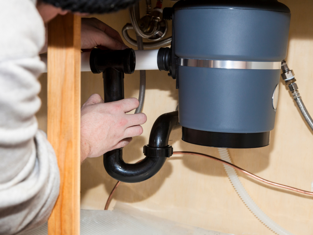 Person installing or repairing plumbing pipes under a kitchen sink, connecting pipes to a garbage disposal unit and ensuring proper alignment for preventing clogs.